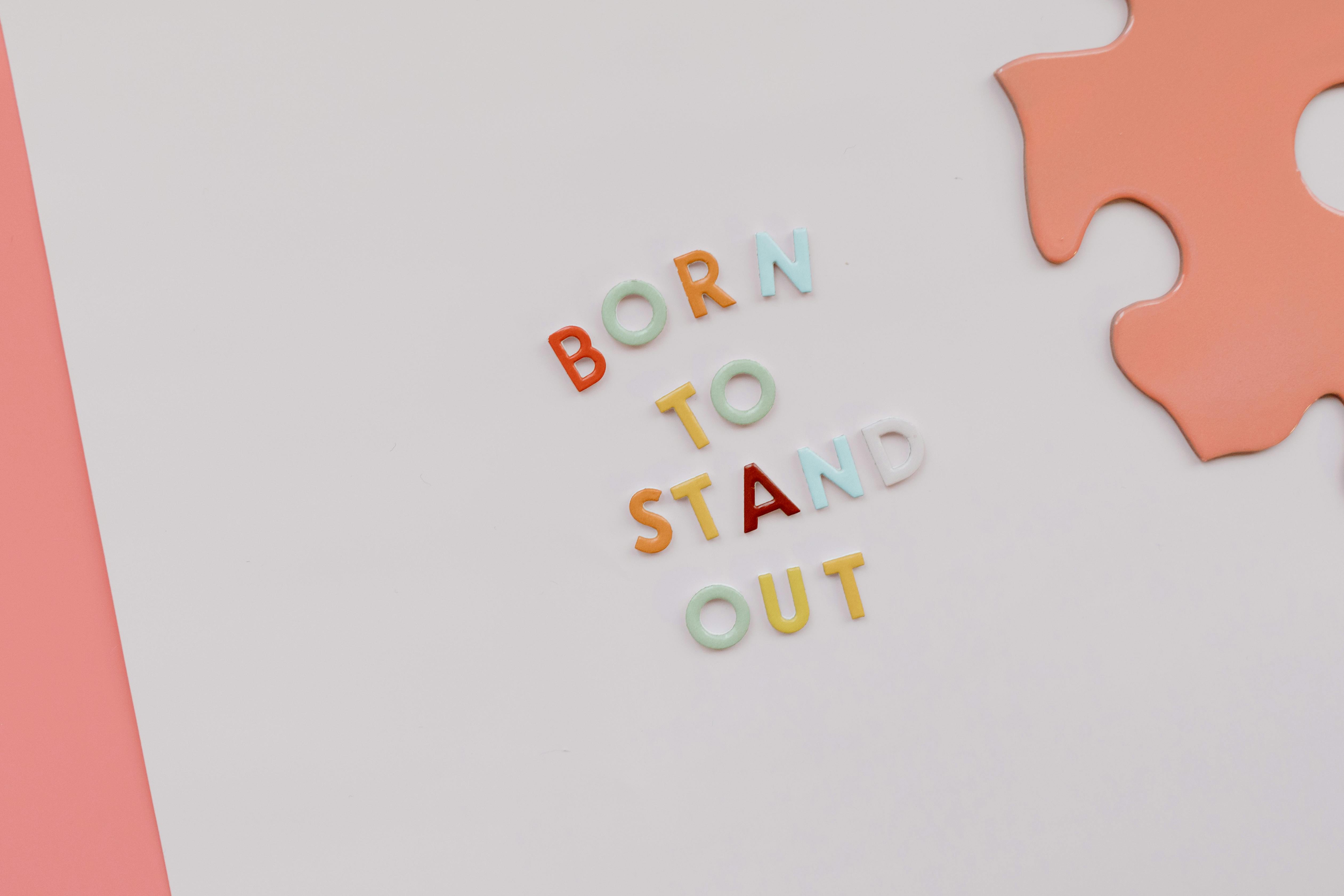 Colourful plastic letters spelling 'BORN TO STAND OUT' on a white surface beside a puzzle piece