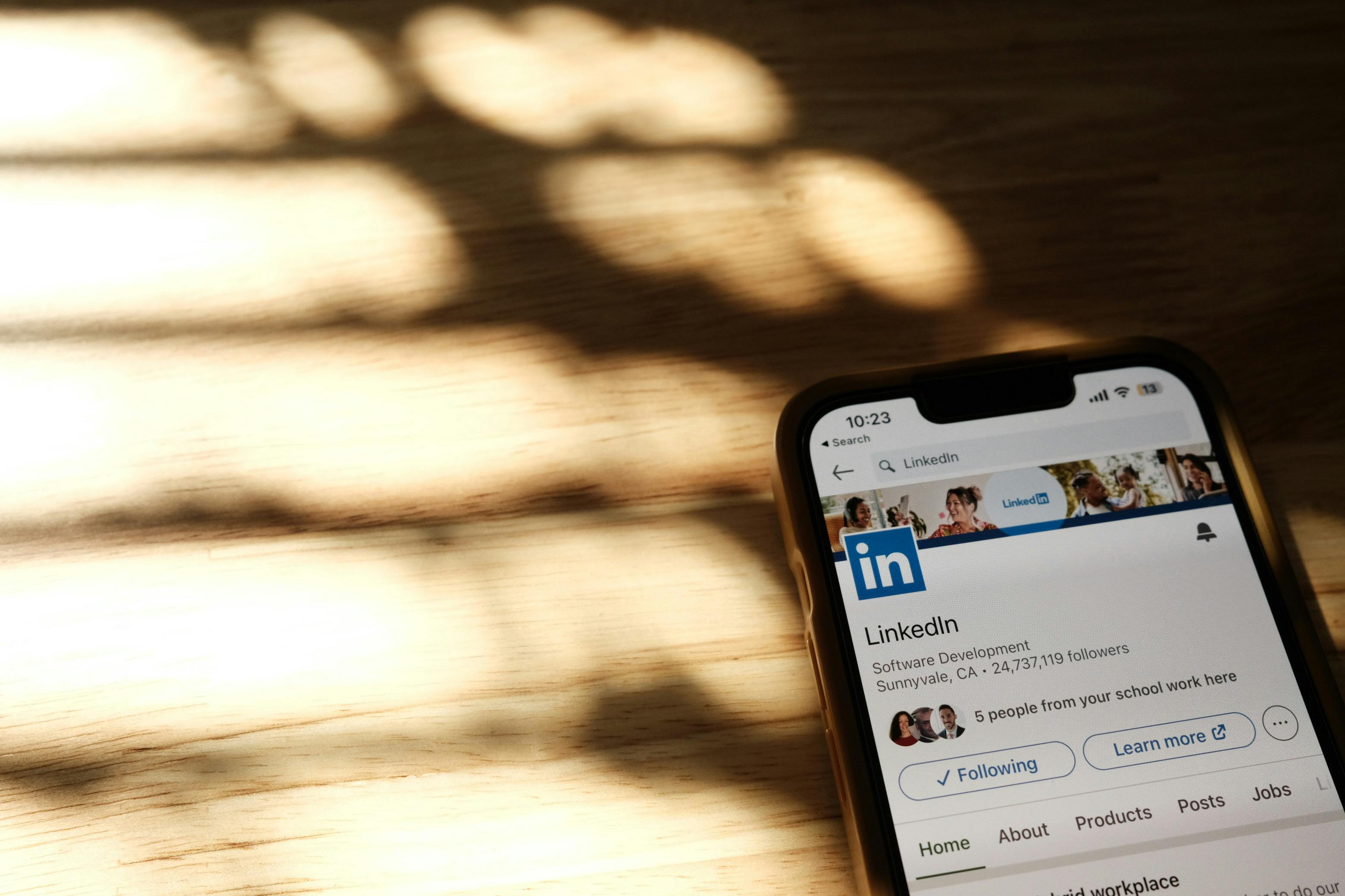A smartphone on a wooden desk displaying a LinkedIn profile, dappled natural light across the surface