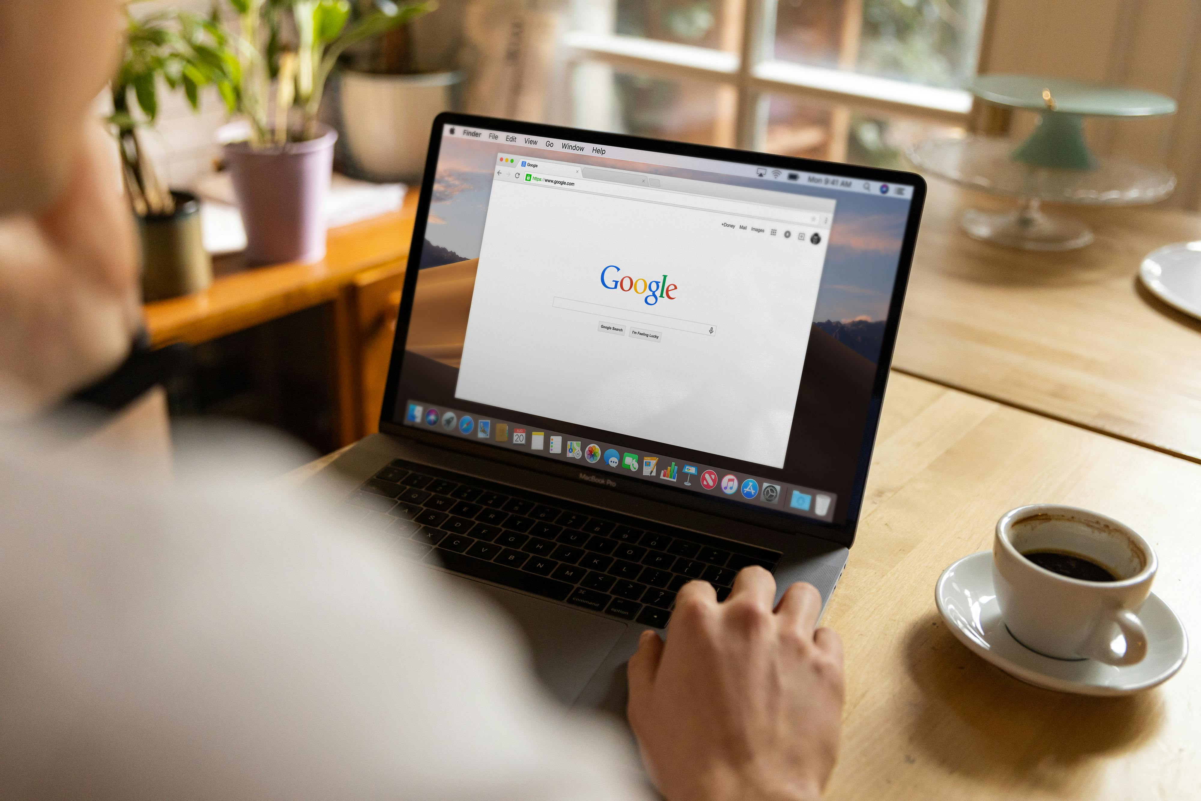 An open MacBook on a wooden desk beside a cup of coffee, homepage visible on screen